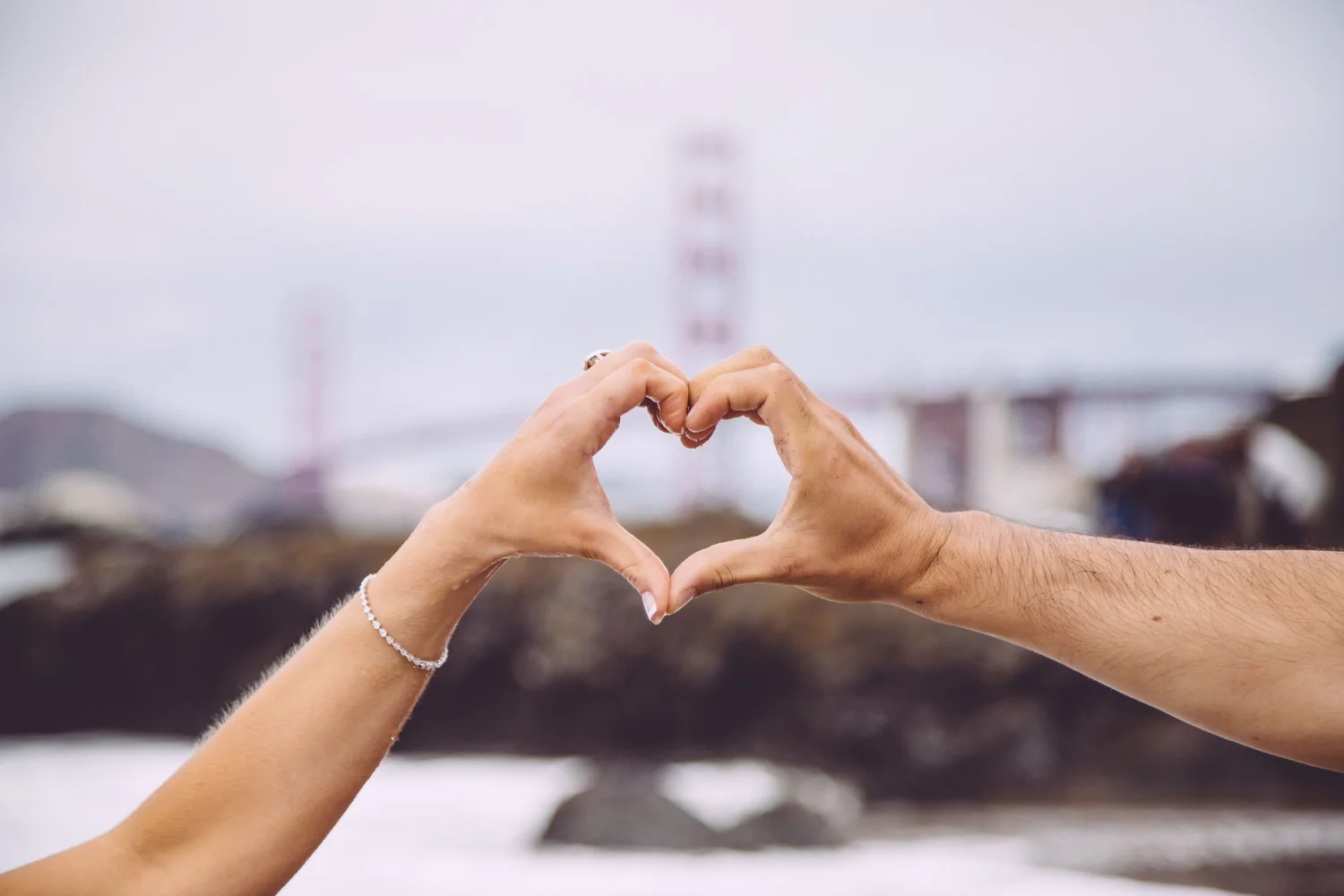 A woman's left arm joins a man's right to form a heart with their thumbs at the base and fingers making the curve. In the background is a bridge, body of water, and rocks