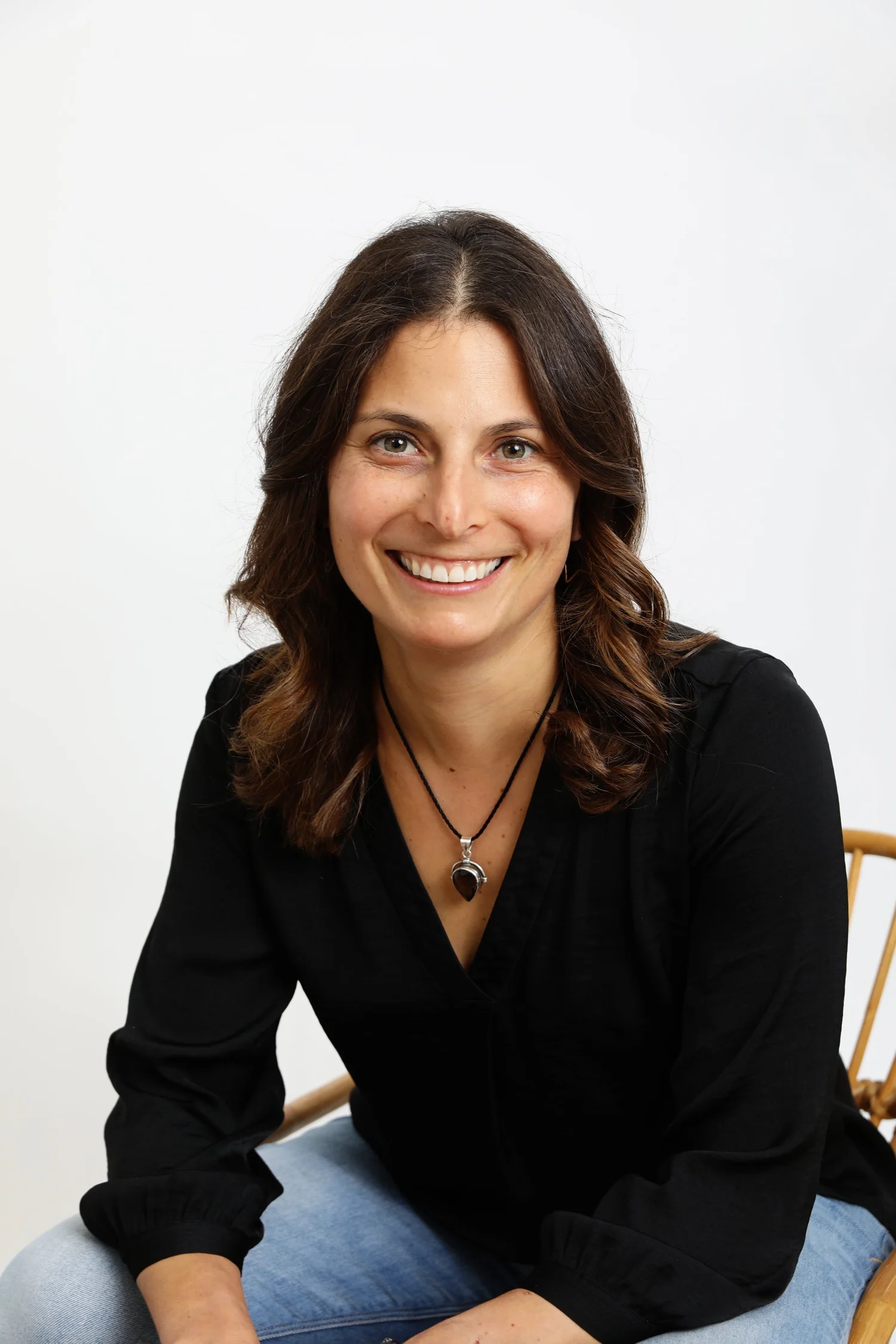 A person with long curly hair sits in a chair, leaning forward toward the camera, with a warm loving smile on their face.