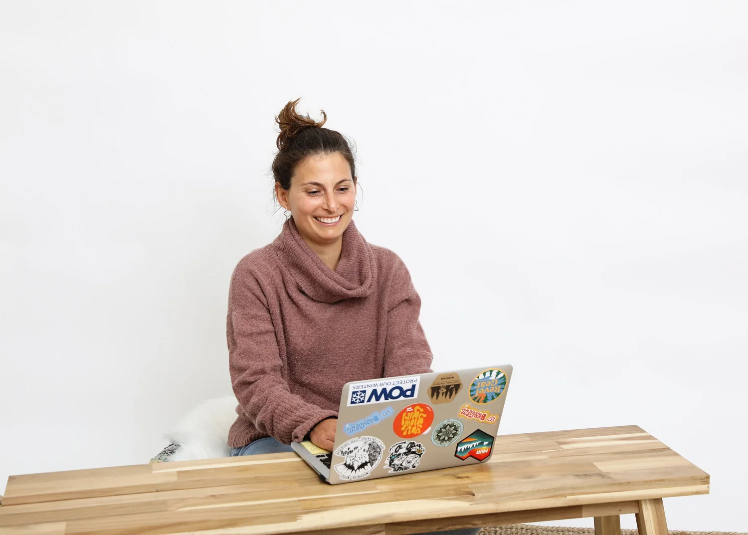 Bara's wears a loose sweater, her hair up in a bun. Her laptop, covered in stickers, sits in front of her. She has a huge warm inviting grin.