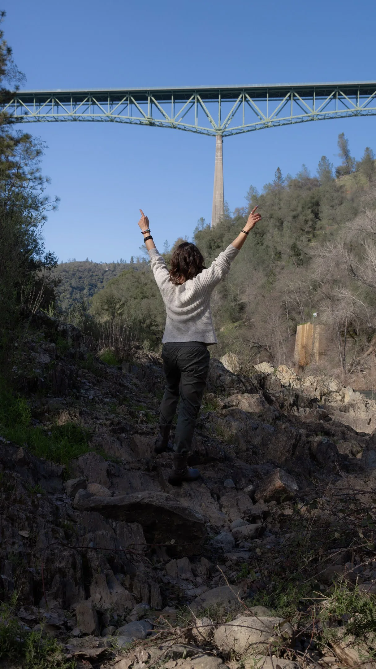 Bara on a rocky riverbank below a high bridge, both arms raised